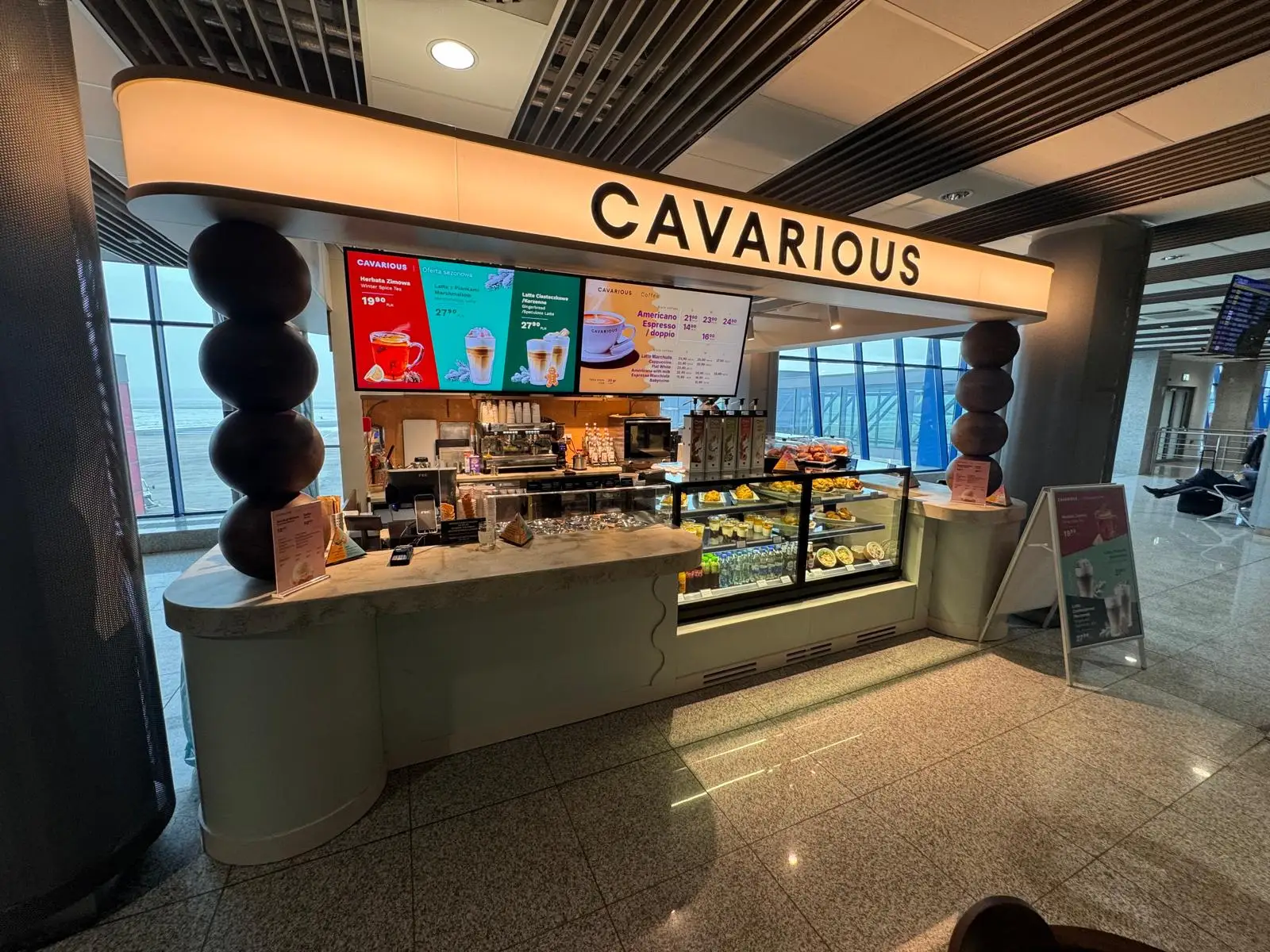 Nowoczesny bar kawowy "CAVARIOUS" w terminalu portowym, z widokiem na menu na ekranach i witrynę z ciastami.